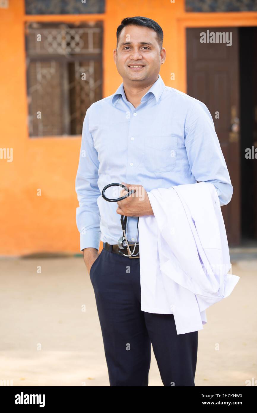 Ritratto di giovane uomo indiano sorridente medico indossando una camicia che tiene stetoscopio e grembiule, sta guardando la macchina fotografica con emozioni positive contro orang Foto Stock