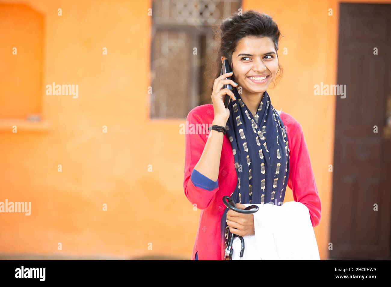 Ritratto di giovane sorridente indiano femmina medico parlare su smartphone o cellulare tenendo stetoscopio e Apron emozioni positive contro la rura arancione Foto Stock