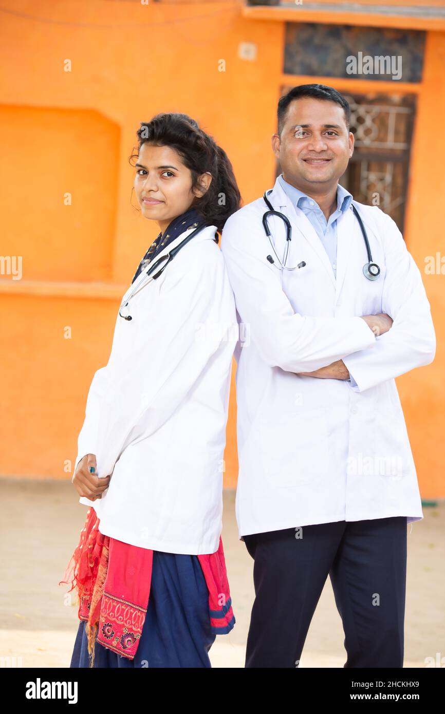 Ritratto di giovane uomo indiano sorridente e femmina medico indossando stetoscopio Croce Arms in piedi guardando fotocamera con emozioni positive contro orang Foto Stock