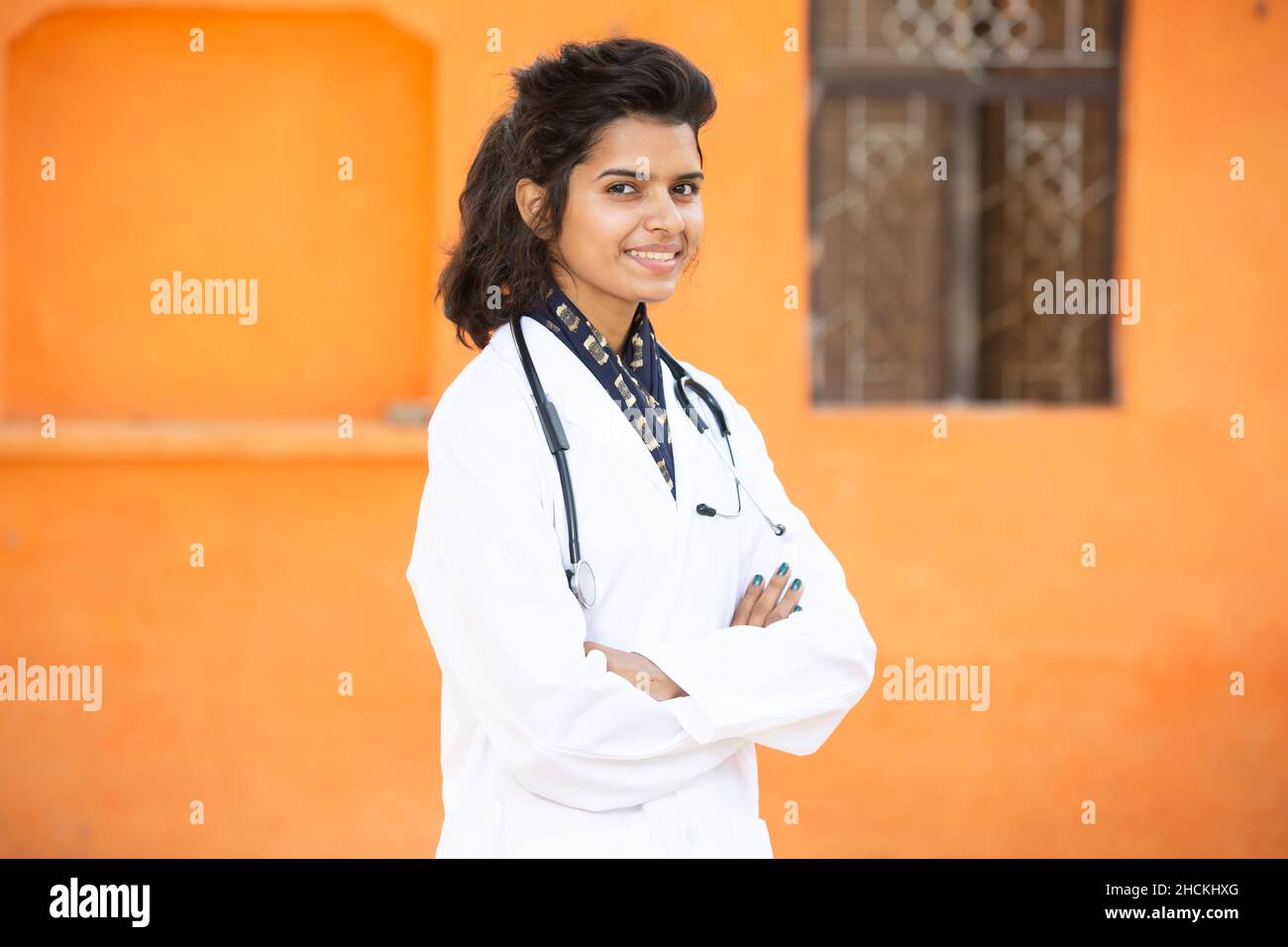 Ritratto di giovane ragazza indiana sorridente medico indossando stetoscopio e grembiule eretto Croce Arms, sta guardando la macchina fotografica con le emozioni positive agai Foto Stock