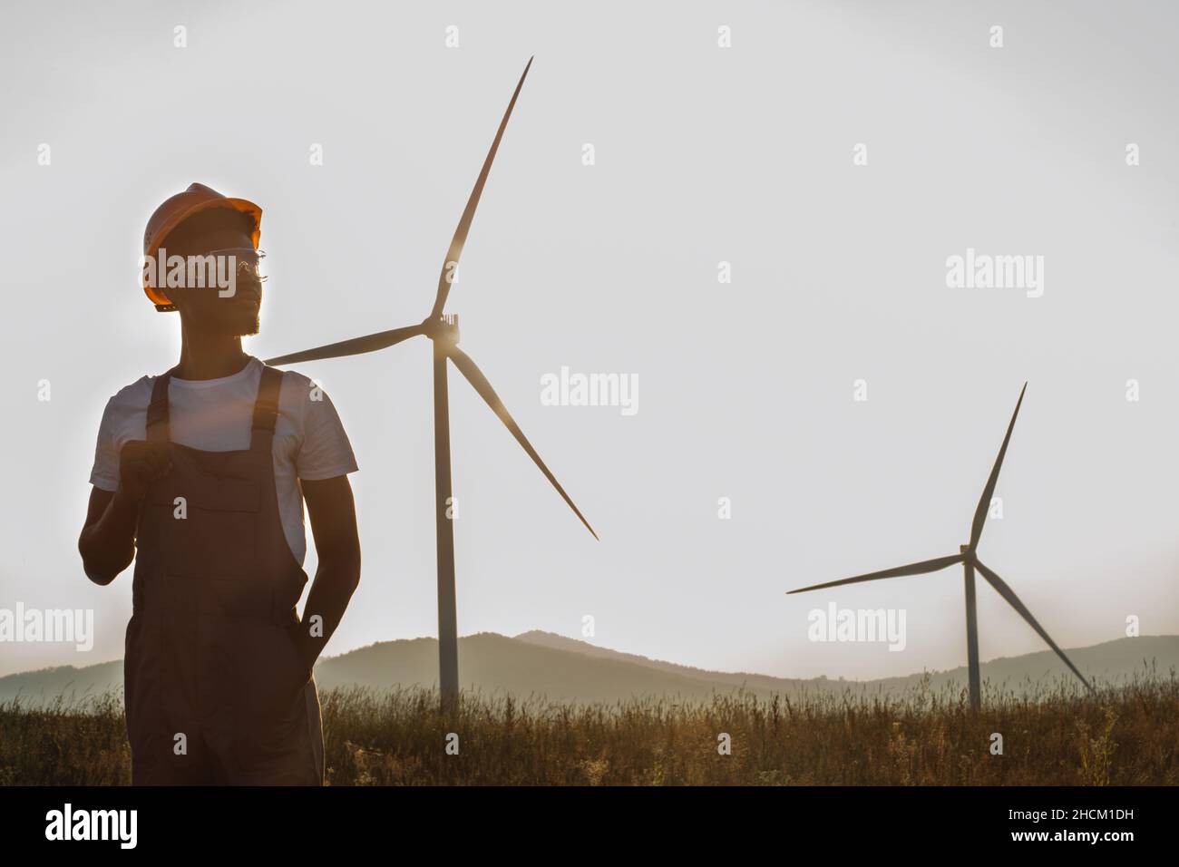 Tecnico afro-americano qualificato in uniforme, casco e occhiali guardando da parte mentre si posa in fattoria con turbine eoliche. Sfondo del tramonto estivo. Concetto di lavoro industriale. Foto Stock