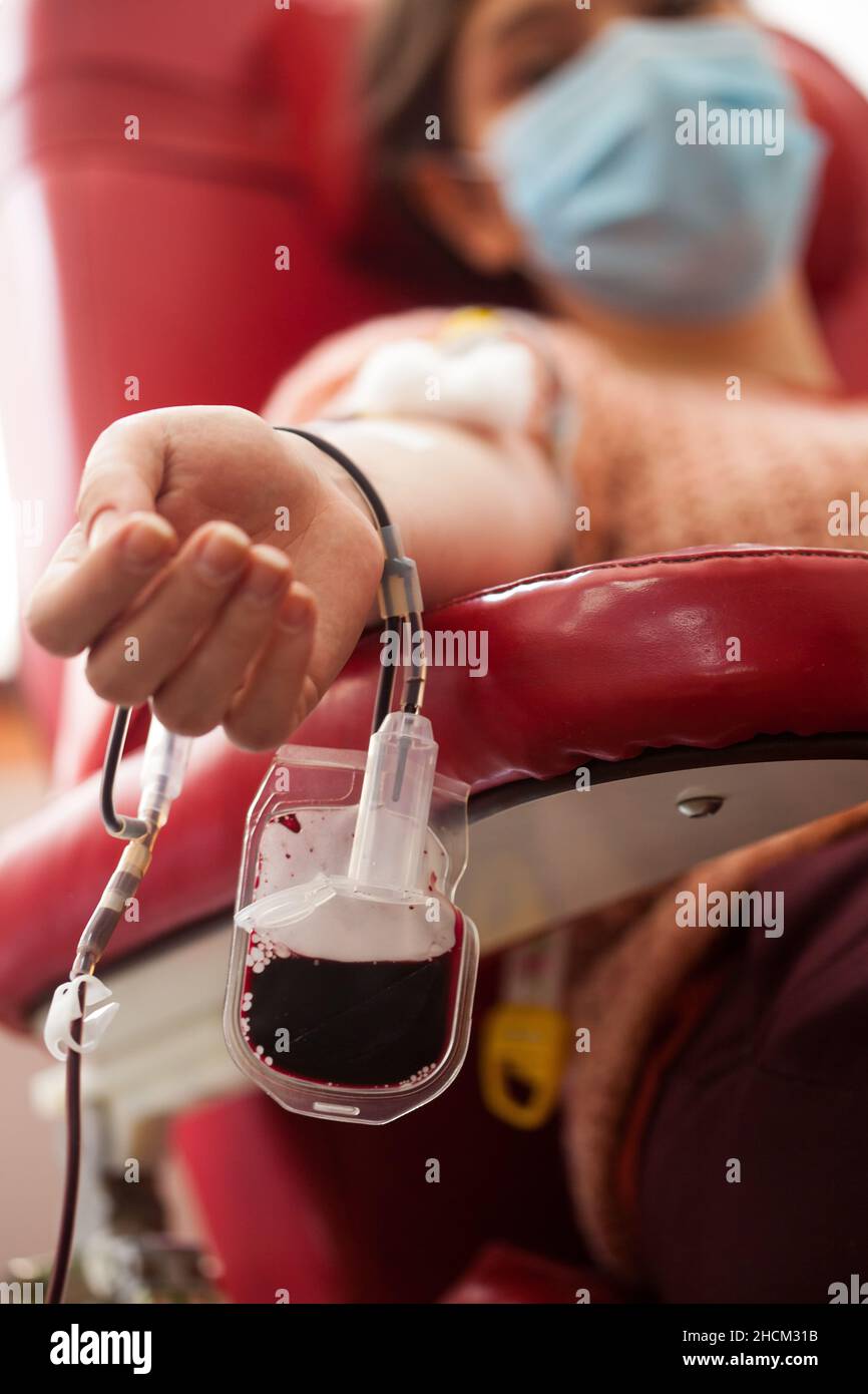 Giovane donna che dona sangue in ospedale o laboratorio, seduta in sedia con sacco di sangue a fuoco, convalescente COVID-19 paziente trasfusione plasma Foto Stock