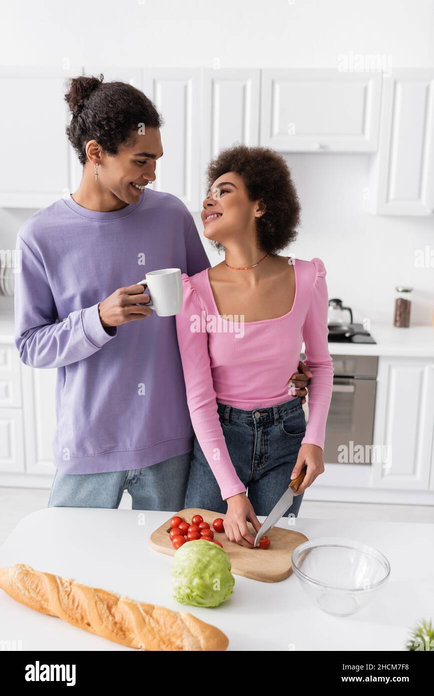 Uomo sorridente che tiene la tazza e abbraccia la ragazza americana africana che taglia il pomodoro di ciliegia a casa Foto Stock