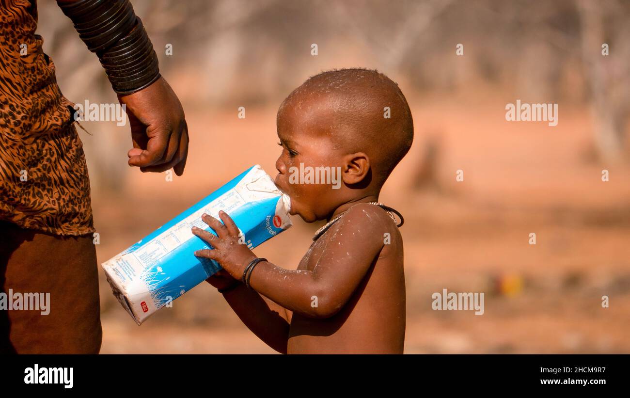 Namibia rurale - 21 agosto 2016. Un ragazzino di un villaggio tribale di Himba beve il latte direttamente dalla scatola mentre si trova accanto alla madre all'aperto. Foto Stock