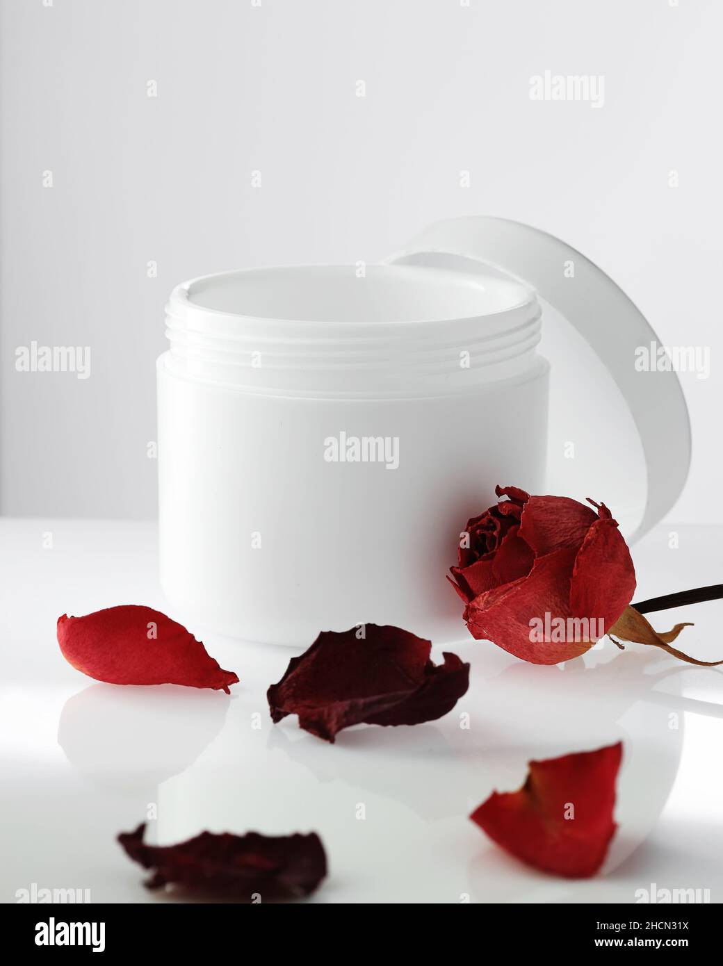 Svuotare il vaso di plastica bianco con il coperchio aperto su sfondo chiaro. Petali di rose rosse. Cosmetici rigeneranti. Prodotto per la cura della pelle del corpo e del viso. Layout per il logo. Cosmetologia e spa. Foto Stock