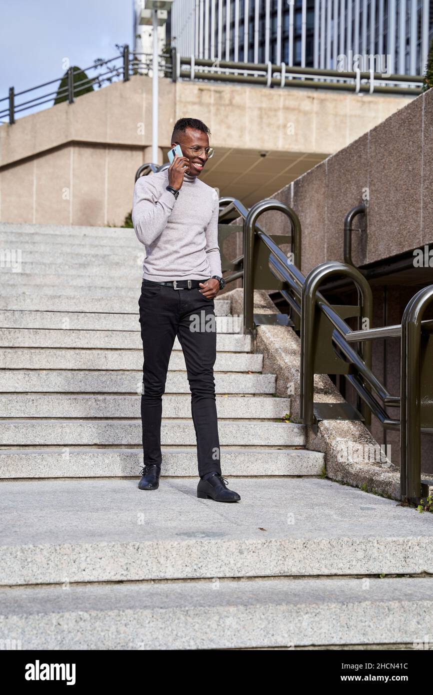 Positivo African American maschio in abiti casual smart tenendo la mano in tasca e rispondere telefono cellulare mentre si cammina su gradini, in giornata di sole in città Foto Stock