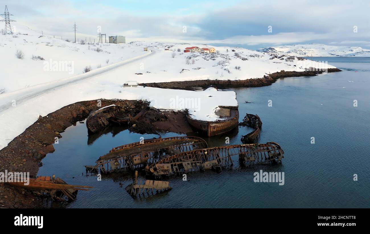 Vista aerea della costa innevata invernale con barche in rovina dopo il naufragio. Vecchie barche da pesca affondate sulla riva del Mare di Barents. Foto Stock