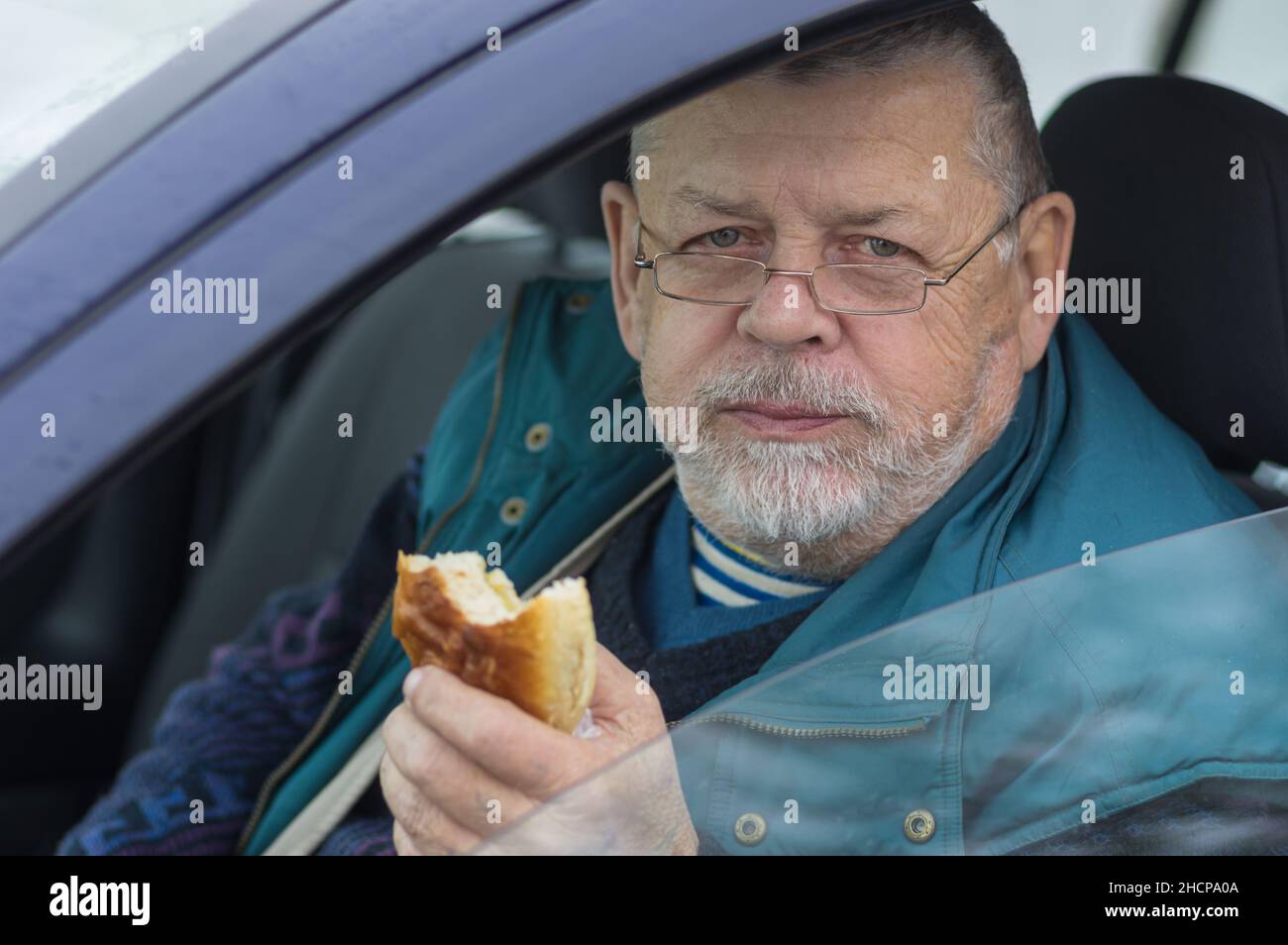 Barbuto caucasico conducente senior prendendo patty e guardando in macchina fotografica mentre si siede all'interno della sua auto Foto Stock