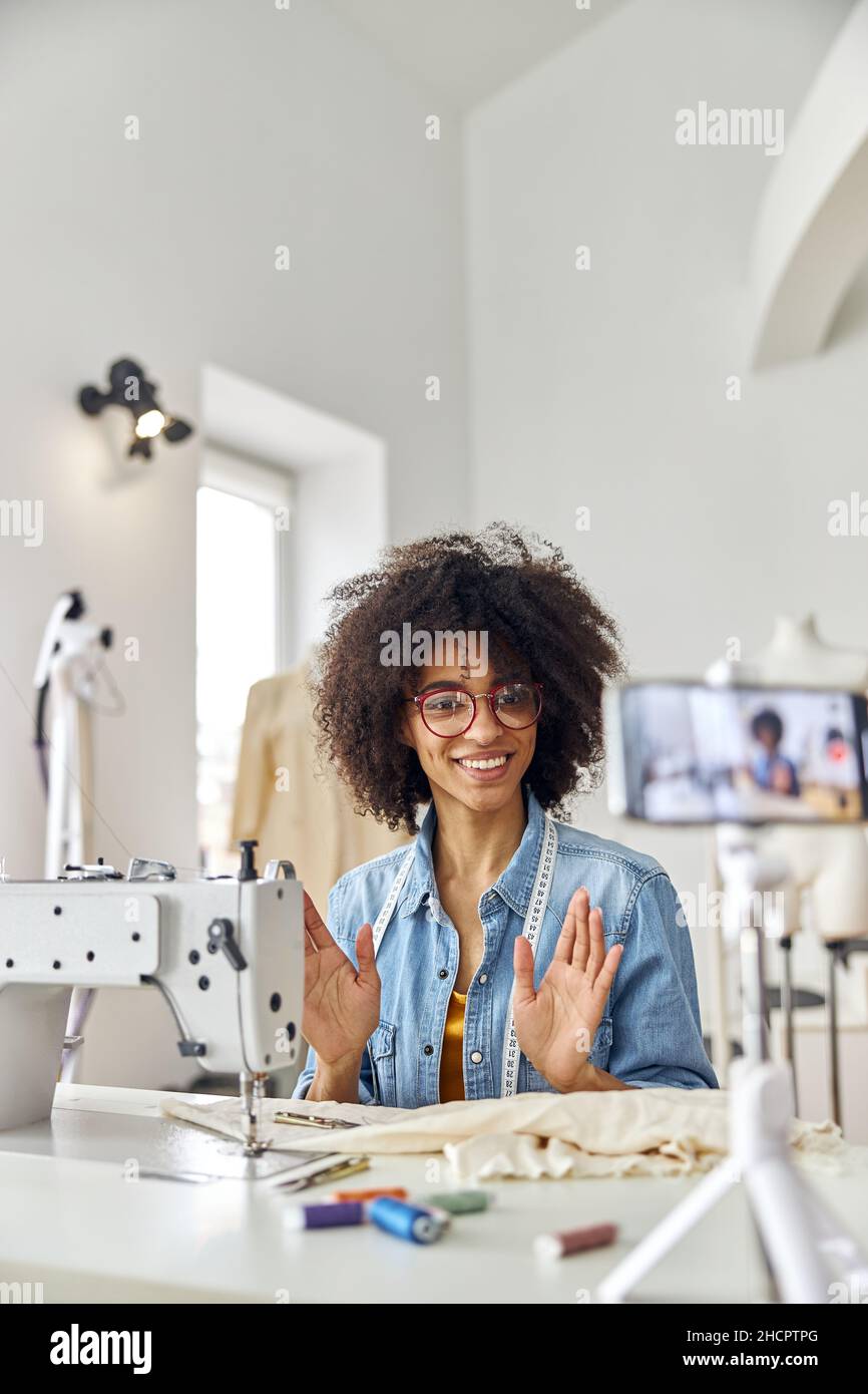 Donna afro-americana dressmaker guarda la macchina fotografica del telefono sul posto di lavoro con la macchina da cucire in studio Foto Stock