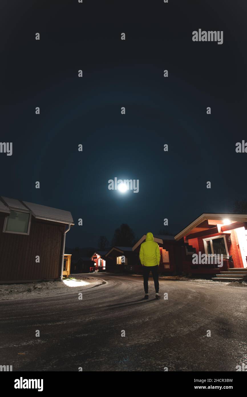 Ambizioso turista in una giacca verde guarda l'effetto alone della luna nella città norvegese settentrionale di Tromso tra le tipiche capanne rosse. Una luna si Foto Stock
