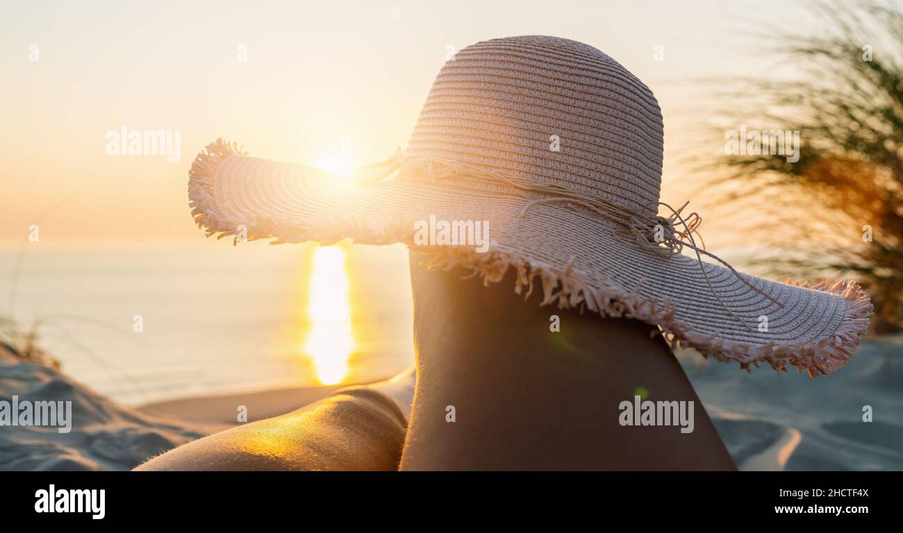 Prospettiva in prima persona delle gambe della donna con cappello di paglia sulla spiaggia al tramonto Foto Stock