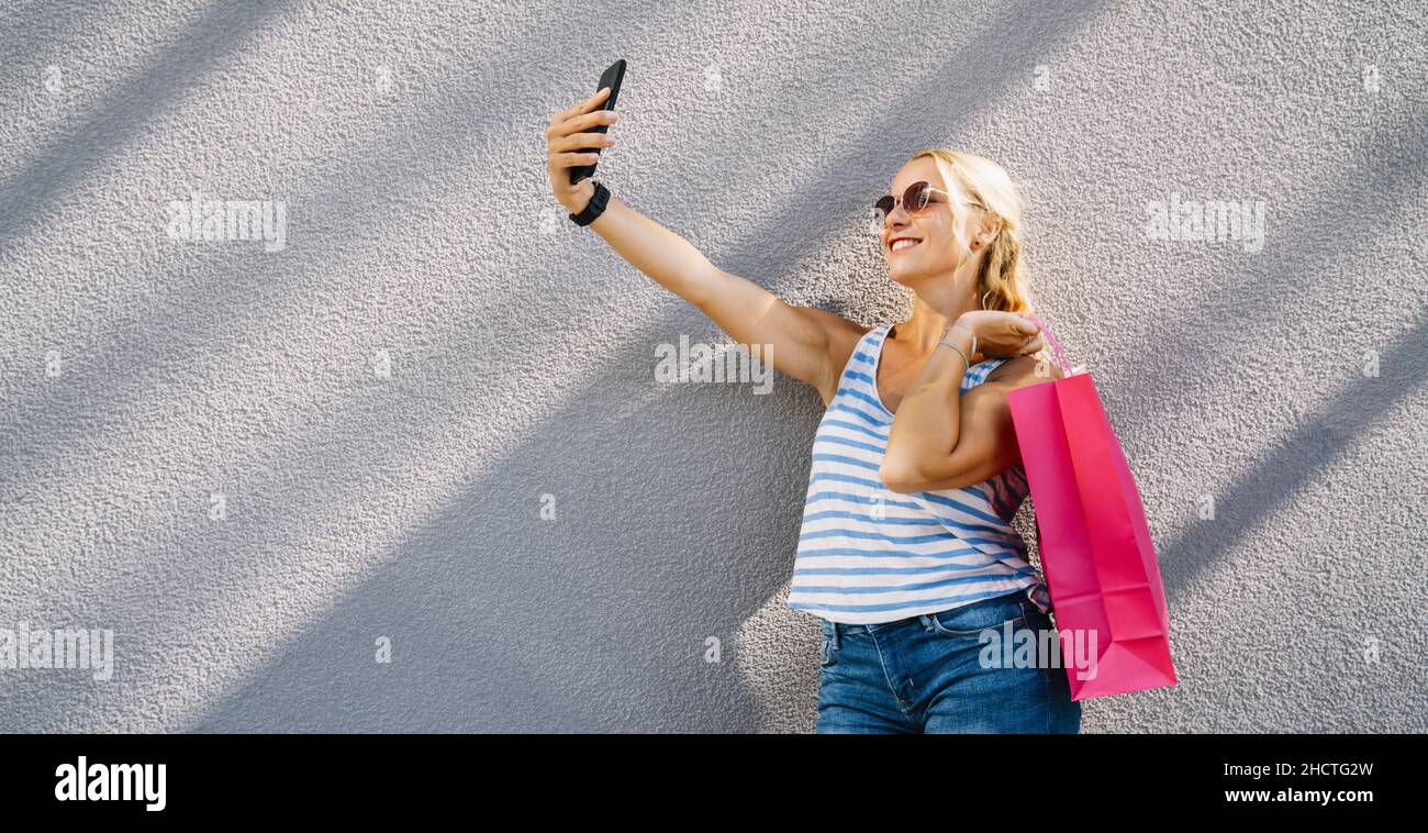 Giovane donna attraente che prende selfie con smartphone con shopping bag dopo lo shopping. Tenere il cellulare e fare selfie. Foto Stock