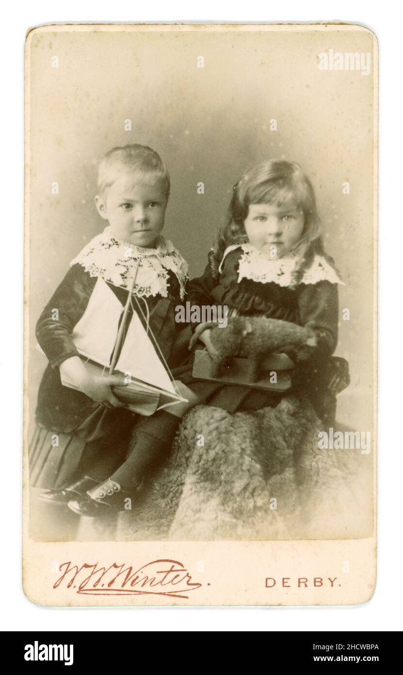 Originale CDV vittoriano di due bambini piccoli - ragazzo con barca giocattolo, ragazza con pony giocattolo -, (o possibilmente un fratello più giovane in abito di una ragazza come era la moda per i ragazzi molto giovani in epoca vittoriana) Questo elaborato stile di abbigliamento per bambini con abito in velluto e collari in pizzo fu influenzato dal popolare romanzo del 1885-1886 "Little Lord Fauntleroy" di Frances Hodgson Burnet. Il ragazzo che regge lo yacht indossa una gonna pieghettata di velluto (anche non raro abbigliamento per ragazzi nel 1880). Questo carte de visite (biglietto da visita) circa fine 1880 - studio di W.W. Winter of Derby, East Midlands, Inghilterra, Regno Unito Foto Stock