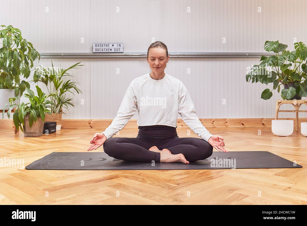 Rilassato barefoot femmina chiudere gli occhi e gesturing Gyan mudra mentre si siede su tappeto in posa di loto e meditating durante la sessione di yoga in studio luce Foto Stock