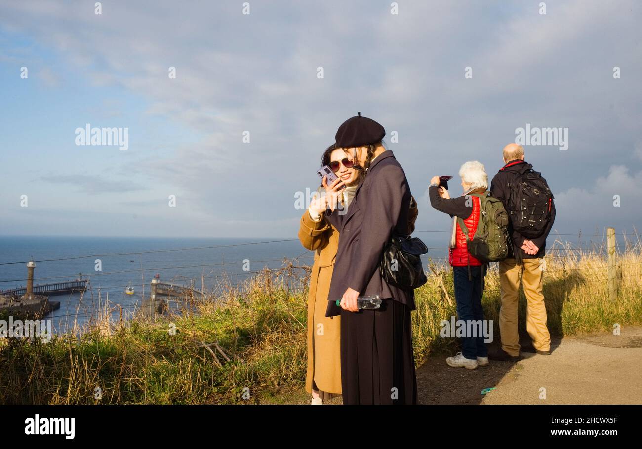 Due giovani donne cinesi che guardano i loro telefoni sopra il porto di Whitby in una giornata di sole invernale Foto Stock