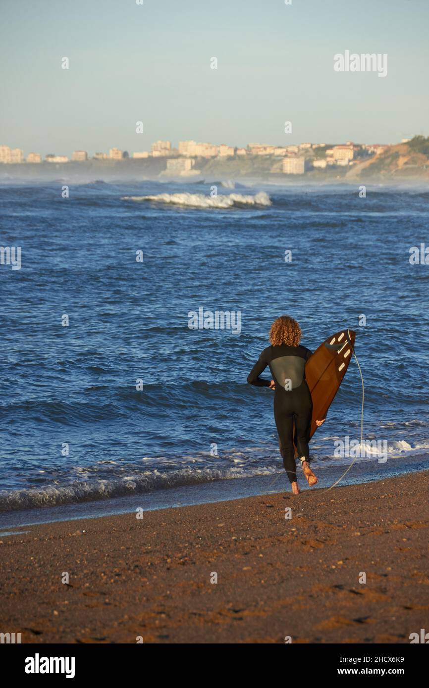 dietro colpo di un surfista riccio che si avvicina al mare con la sua tavola in mano, si possono vedere le onde e la scogliera con le case sullo sfondo Foto Stock