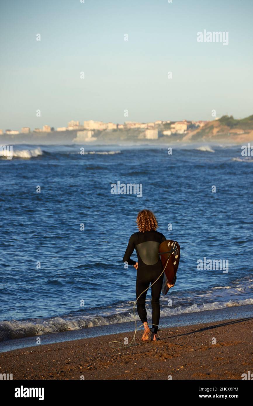 dietro colpo di un surfista riccio che si avvicina al mare con la sua tavola in mano, si possono vedere le onde e la scogliera con le case sullo sfondo Foto Stock