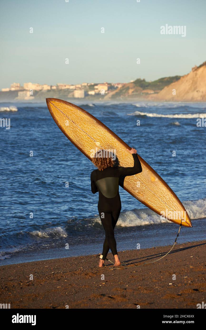 dietro colpo di un surfista riccio che si avvicina al mare con la sua tavola in mano, si possono vedere le onde e la scogliera con le case sullo sfondo Foto Stock
