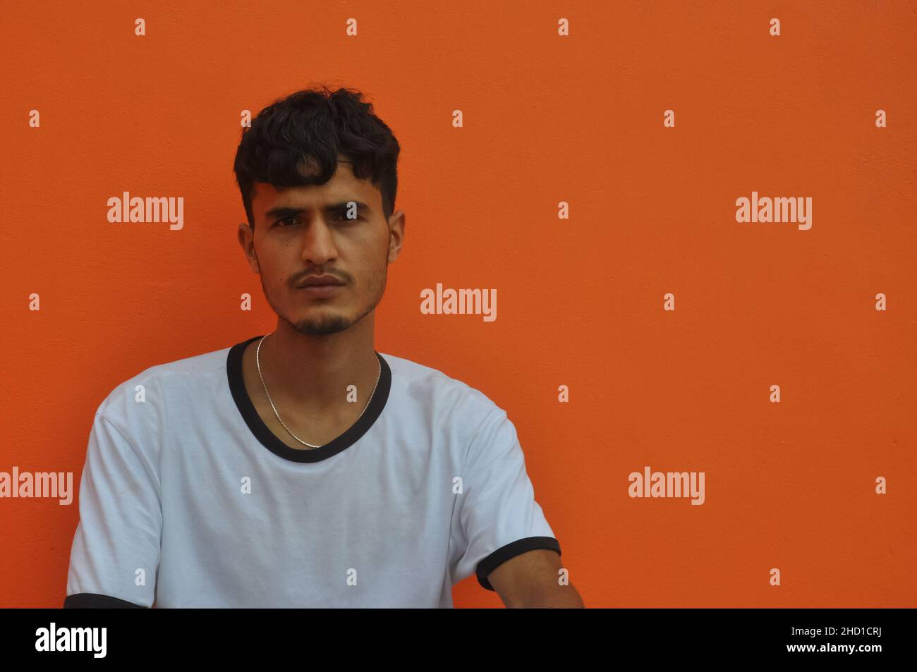Vista frontale di un ragazzo dell'Asia meridionale che guarda la macchina fotografica, seduto su sfondo arancione della parete con spazio copia Foto Stock