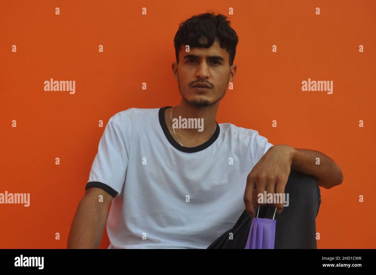 Vista frontale di un ragazzo giovane che guarda la macchina fotografica con la maschera protettiva per il viso in mano, seduto su sfondo arancione parete con spazio copia Foto Stock