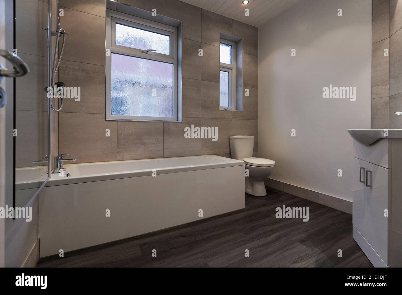 Bagno moderno con vanity unit, grandi piastrelle bianche Barrington, pavimento in vinile LVT grigio e pareti bianche, vasca doccia, schermo doccia e toilet Foto Stock