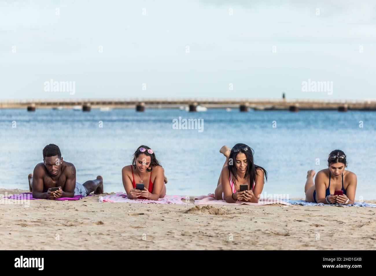 Diversi uomini e donne che si trovano su coperte e navigare telefoni cellulari mentre si riposa sulla spiaggia sabbiosa vicino al mare sull'isola di Lanzarote, Spagna Foto Stock