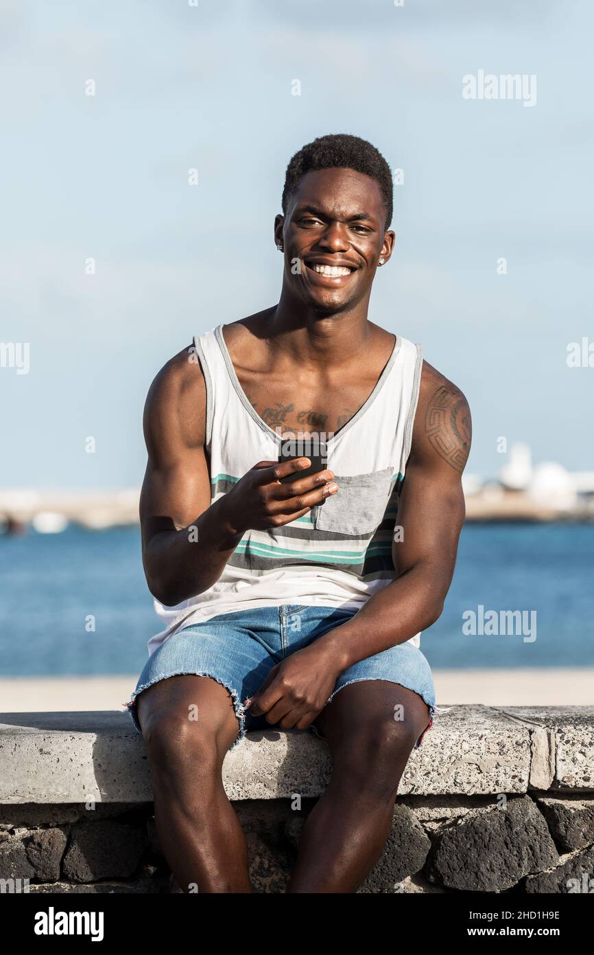 Happy maschio nero in estate vestiti seduti su pietra recinzione contro il mare a Lanzarote e navigare smartphone mentre si gode la giornata estiva e guardando ca Foto Stock