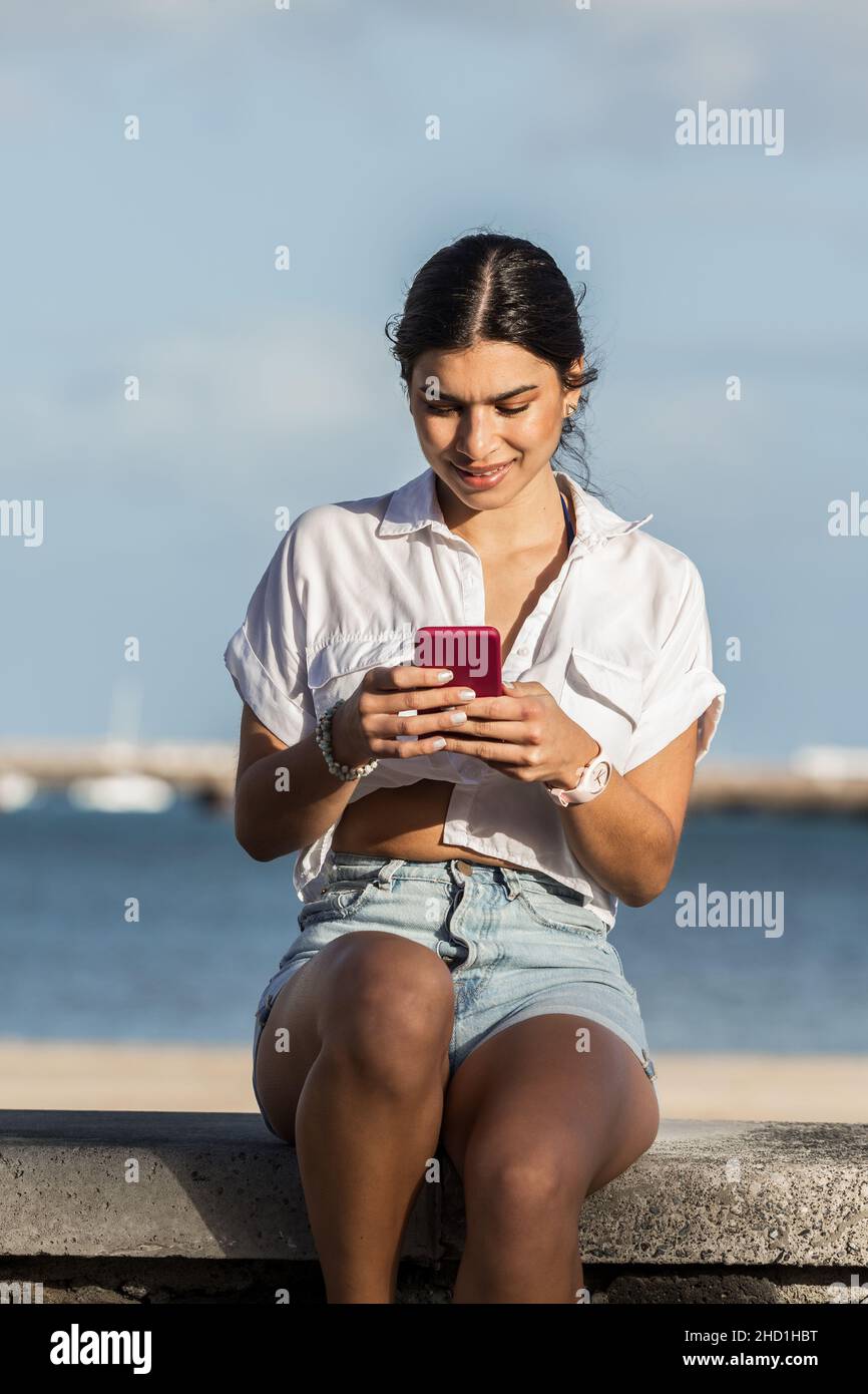 Donna serena seduta sulla recinzione in pietra sul lungomare e navigare sul telefono cellulare mentre si passa il tempo in estate a Lanzarote Foto Stock