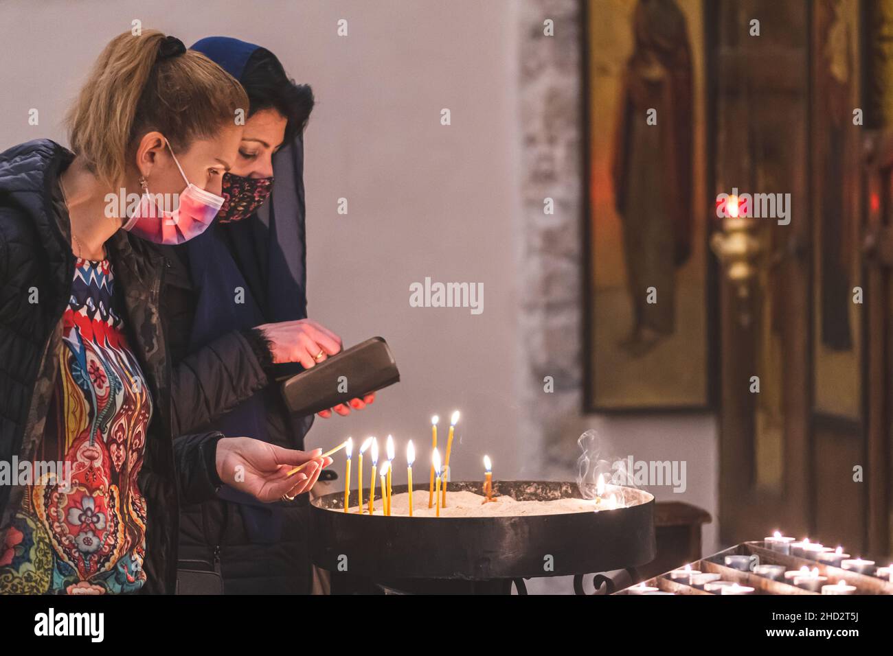 Due belle ragazze caucasiche o donne che indossano maschere e illuminazione candele votive in una chiesa Foto Stock