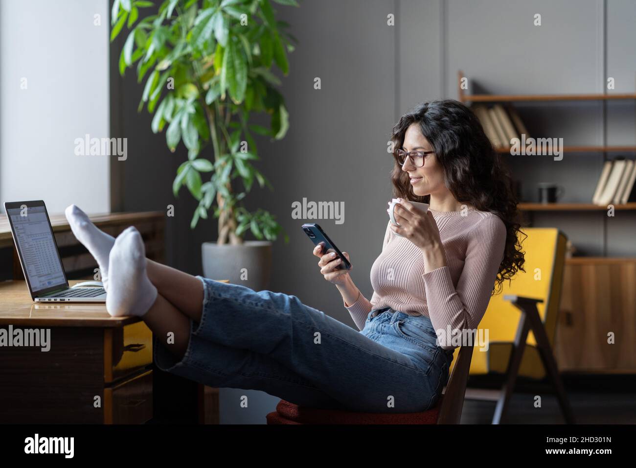 Donna d'affari rilassata bevendo caffè, leggendo le notizie online sullo smartphone mentre si rilassa sul posto di lavoro Foto Stock