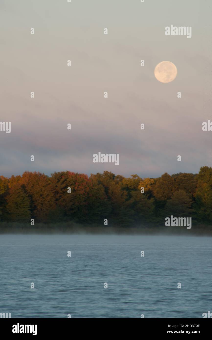 La luna piena raccolto attraverso sottili nuvole su un lago con colori autunnali sulla riva lontana. Foto Stock