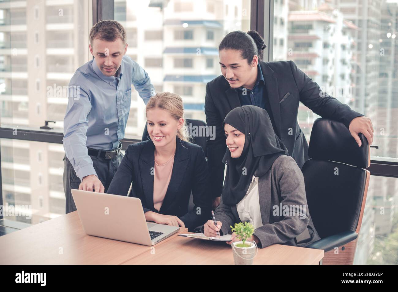 Gruppo di impiegati che lavorano in team lavorando e guardando i notebook insieme ai sorrisi in ufficio, avendo edifici urbani in background. Foto Stock