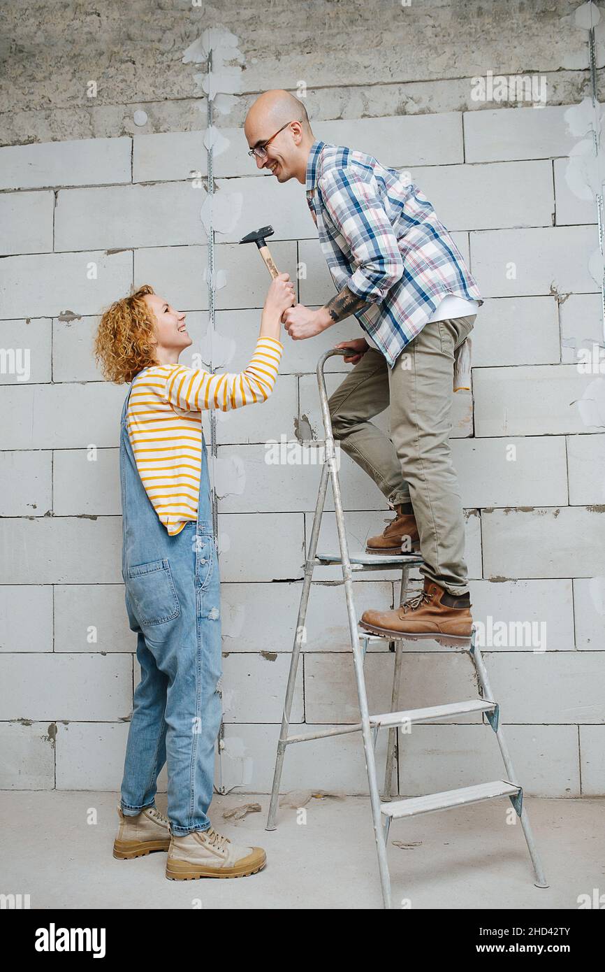 Guardando l'altra coppia facendo nuova revisione appartamento insieme. L'uomo sta salendo su una scala a cavalletto, la donna gli sta passando un martello. Foto Stock