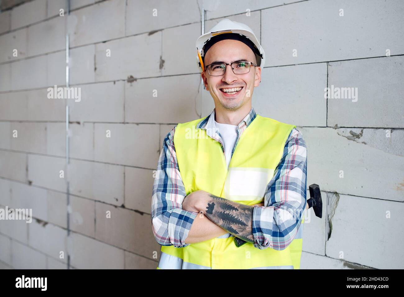 Ritratto di un ingegnere edile in un casco bianco con le mani ripiegate sul petto. Su un muro di mattoni a livello in un edificio incompiuto. Foto Stock