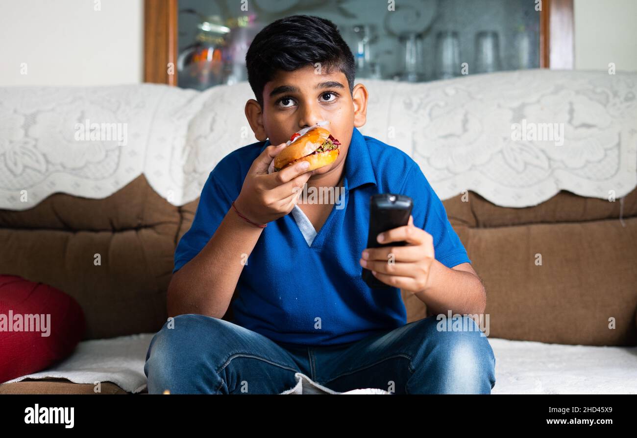 bambino indiano teenger guardando la tv o la televisione mangiando cibo malsano a casa - concetto di attività di lesieur, perdita di tempo e uno stile di vita malsano. Foto Stock
