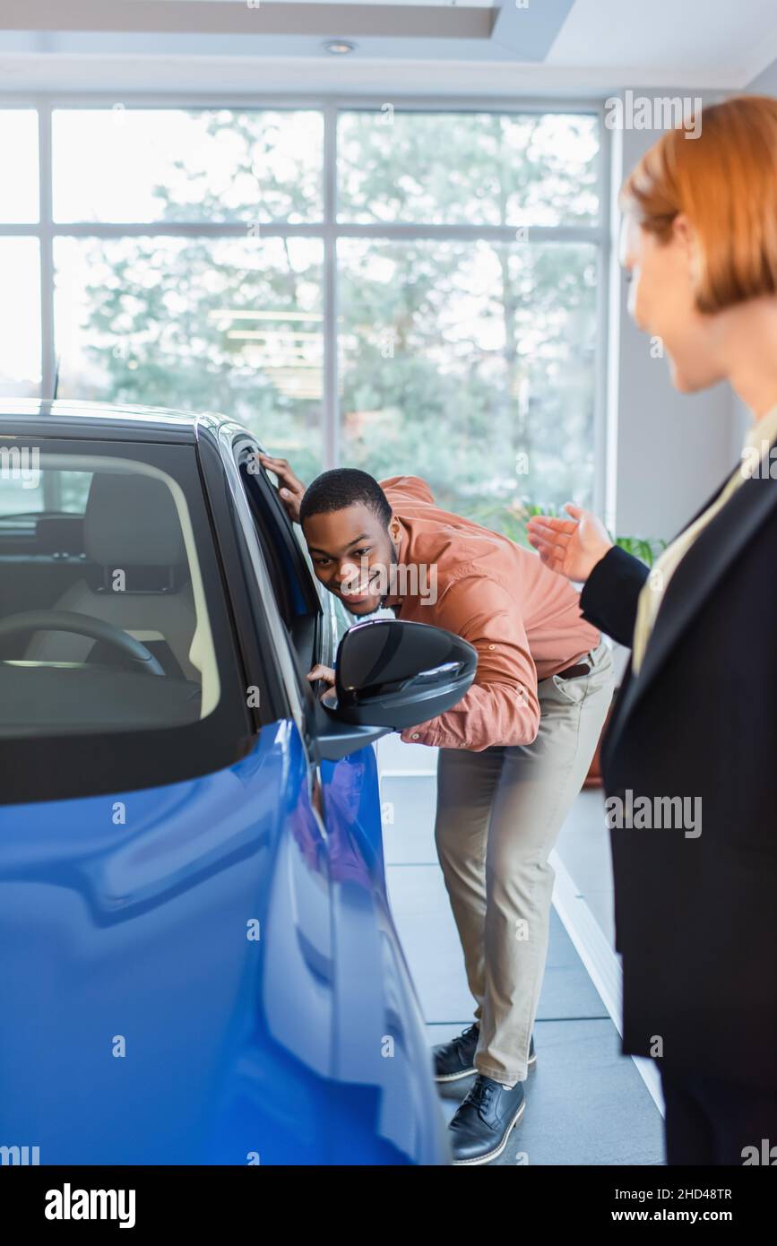 murled concessionario auto che punta con mano vicino felice africano americano uomo guardando all'interno della macchina in showroom Foto Stock