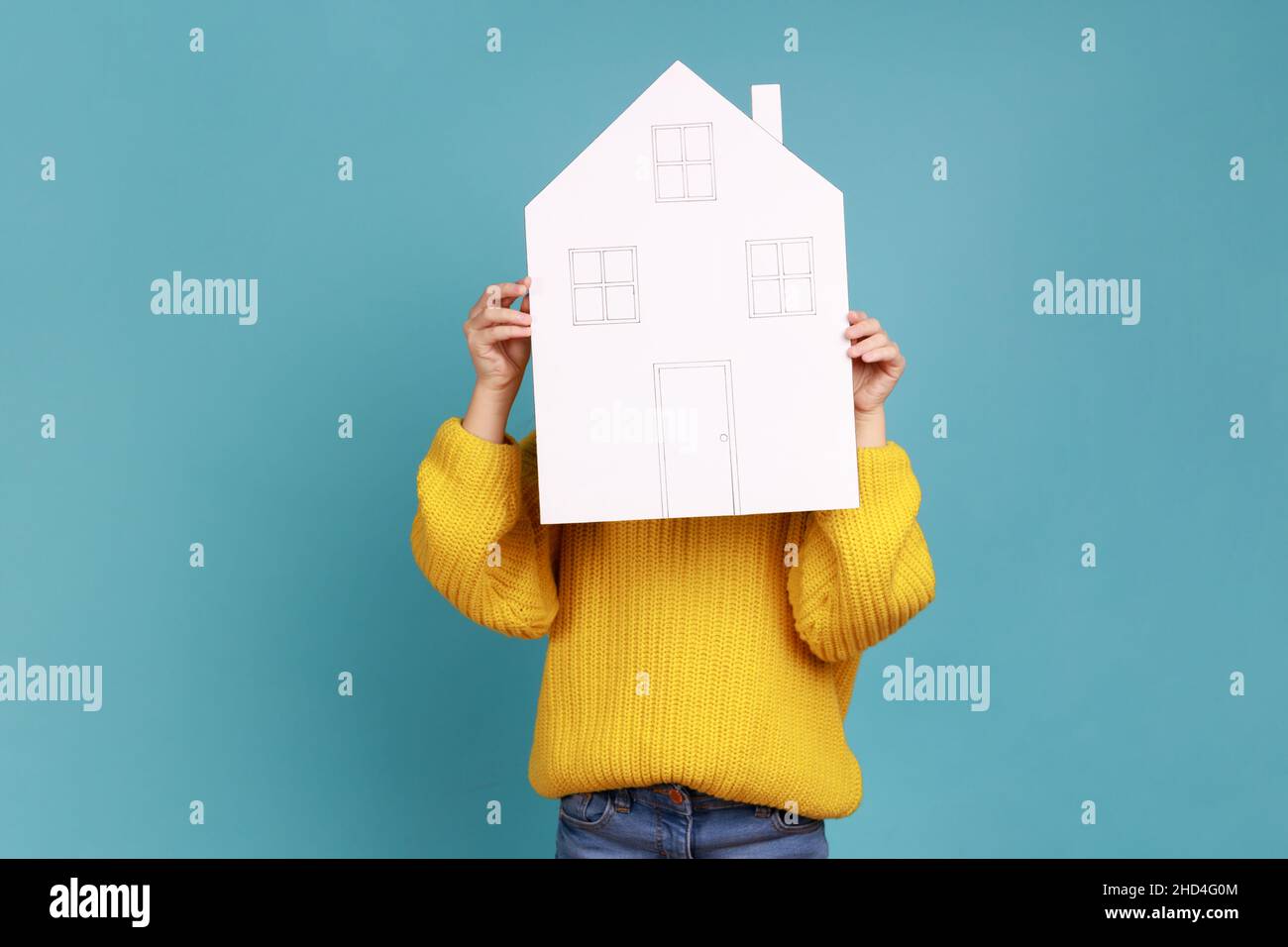 Ritratto di sconosciuto bambino nascosto dietro la casa di carta, abbordabile programma di alloggiamento, pubblicità, indossare giallo casual stile maglione. Studio interno girato isolato su sfondo blu. Foto Stock