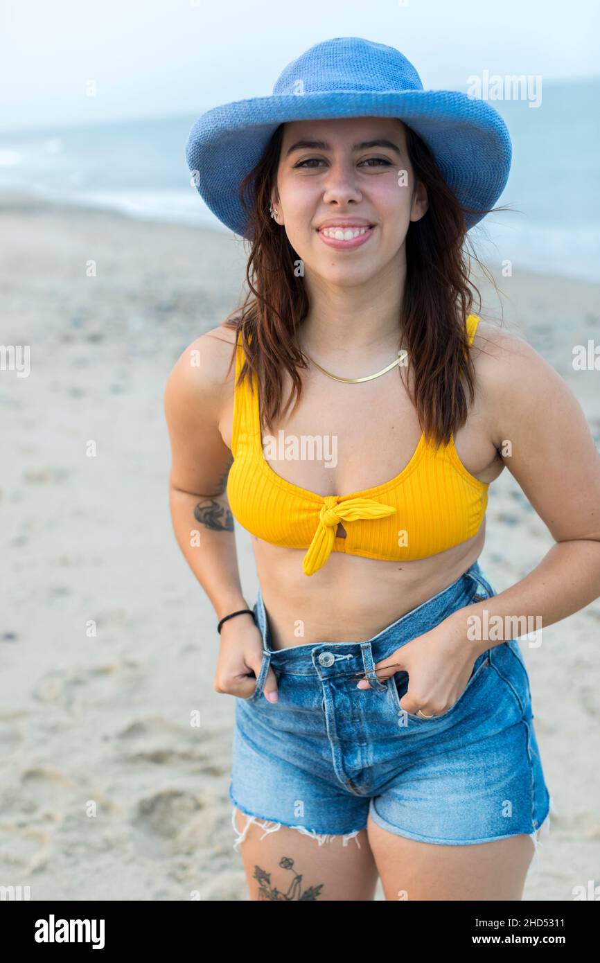 Ritratto di giovane donna in spiaggia in cappello blu, pantaloncini e costume da bagno Foto Stock
