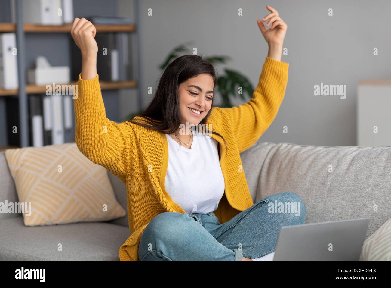 Signora araba molto gioiosa seduta sul divano con computer portatile, gesturando sì, festeggiando il successo a casa, spazio libero Foto Stock