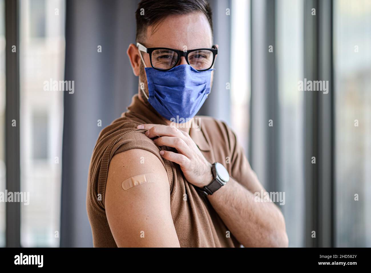 Giovane uomo adulto che mostra puntando al bendaggio sulla spalla del suo braccio indossare maschera protettiva fatto con la vaccinazione. Il maschio è appena stato vaccinato contro Covid1 Foto Stock