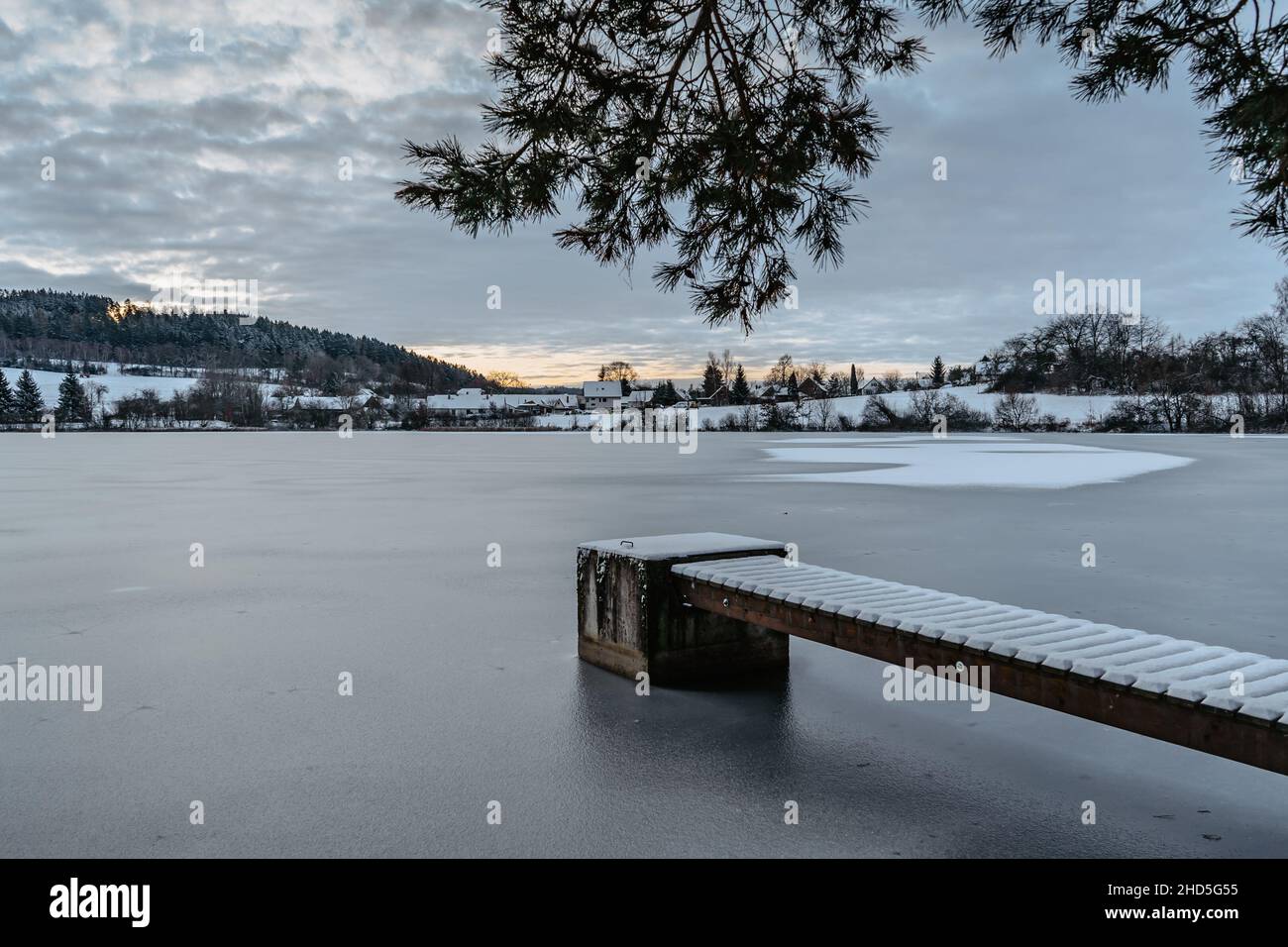 Molo di legno sul lago con neve fresca. Stagno di inverno con piccolo molo all'alba, villaggio in backround.Frosty paesaggio calmo. Bianco inverno campagna nevoso Foto Stock