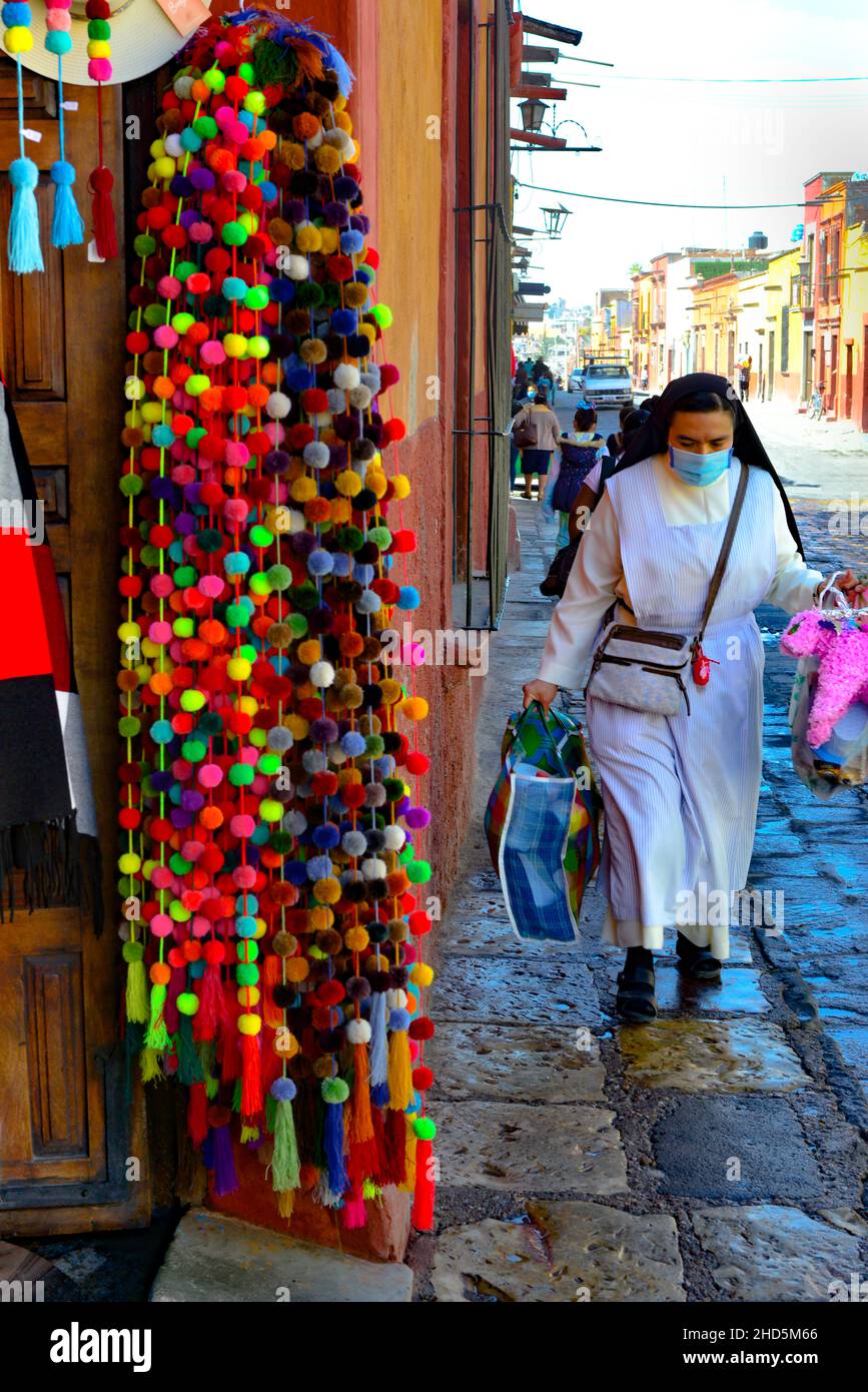 Storefront con prodotti colorati appesi lungo la porta aperta per la strada e marciapiede con una suora in sua abitudine con piñata a San Miguel de Allende, MX Foto Stock