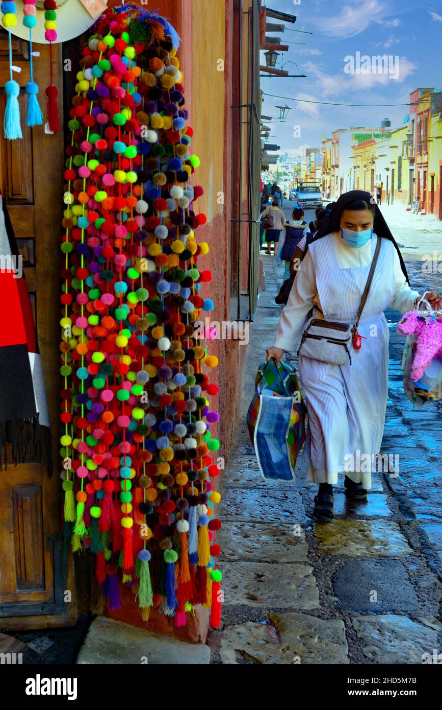 Storefront con prodotti colorati appesi lungo la porta aperta per la strada e marciapiede con una suora in sua abitudine con piñata a San Miguel de Allende, MX Foto Stock