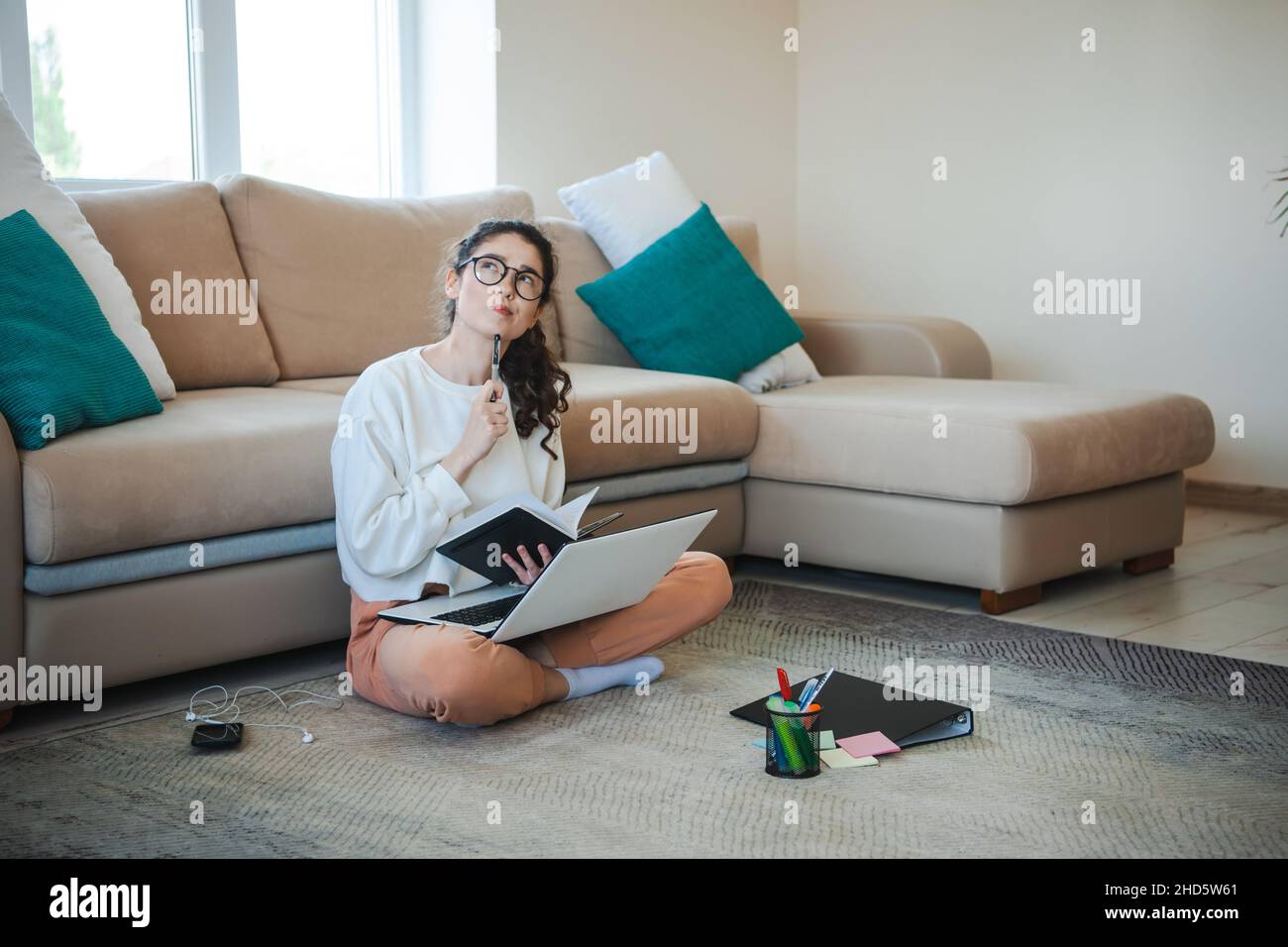 Donna che studia a casa con gambe incrociate sul pavimento appoggiate su un divano che lavora con un computer portatile e note di classe. Donna caucasica che guarda in su mentre Foto Stock