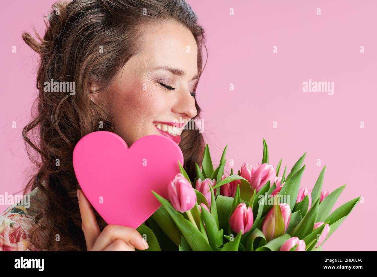 felice giovane donna in abito floreale con bouquet tulipani e cuore rosa su sfondo rosa. Foto Stock