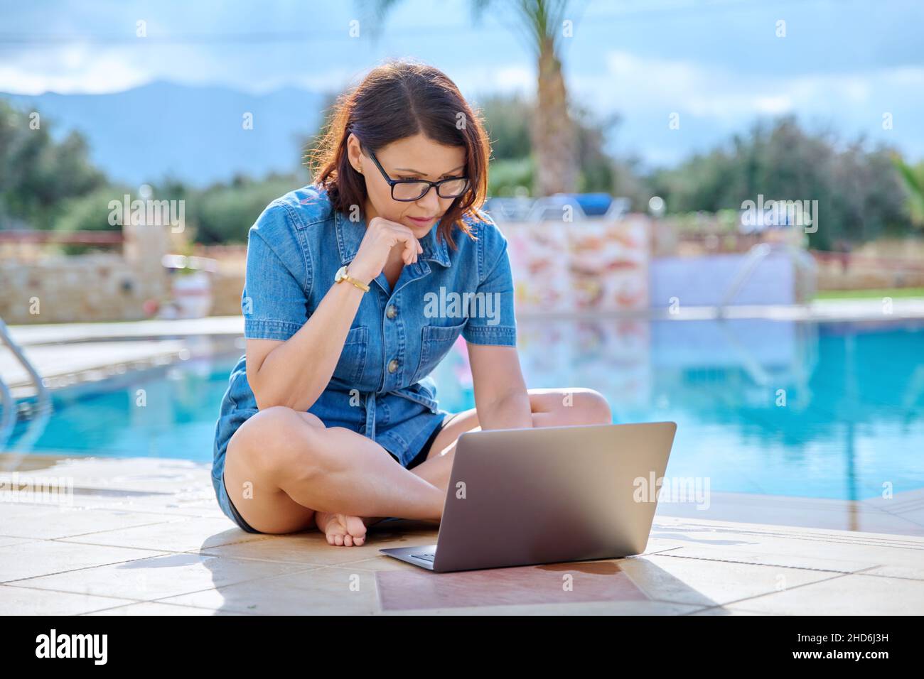 Donna di mezza età che si rilassa vicino alla piscina utilizzando un computer portatile. Foto Stock