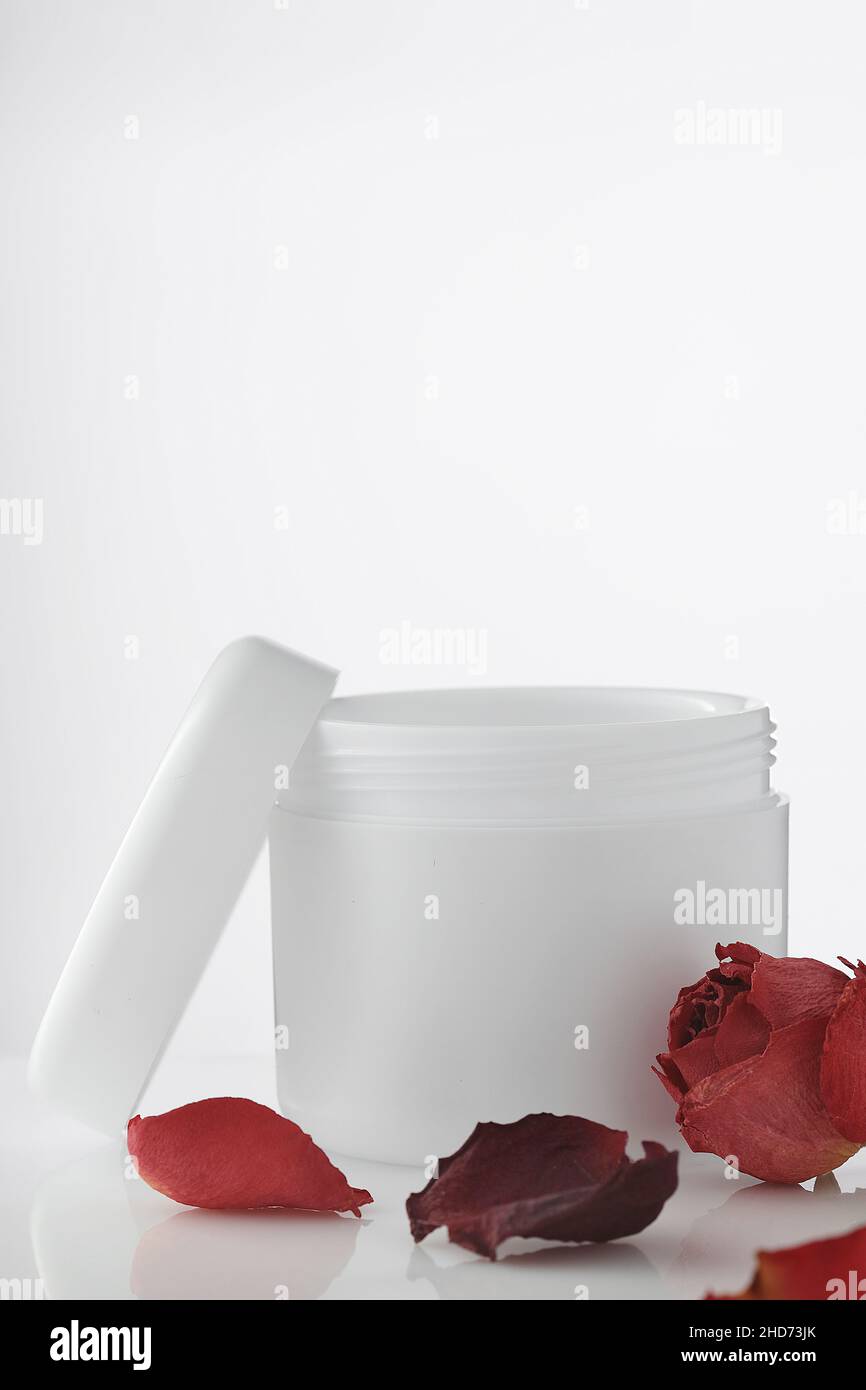 Svuotare il vaso di plastica bianco con il coperchio aperto su sfondo chiaro. Petali di rose rosse. Cosmetici rigeneranti. Prodotto per la cura della pelle del corpo e del viso. Layout per il logo. Cosmetologia e spa. Foto Stock