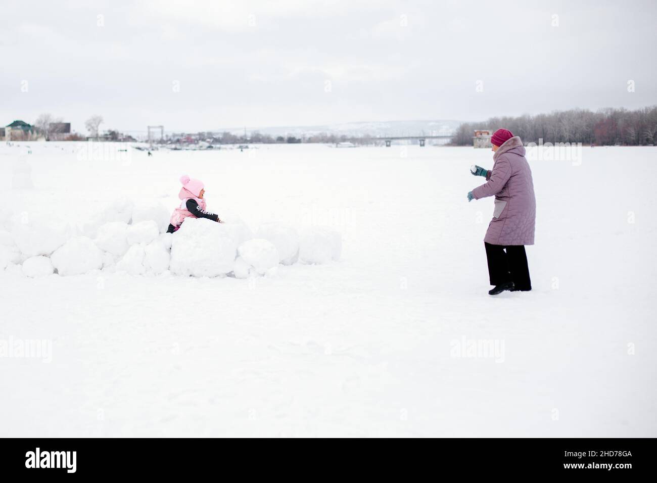 Foto a distanza di bambini che giocano a palle di neve con nonna che si divertono indossando abiti rosa inverno. Sfondo sorprendente pieno di bianco e neve Foto Stock