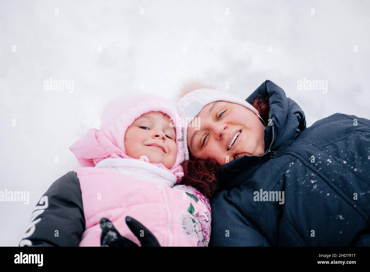 Primo piano di foto di bambino sdraiato nella neve con la madre ed entrambi guardando in macchina fotografica che indossa i vestiti rosa di inverno nella foresta. Sfondo sorprendente pieno di Foto Stock