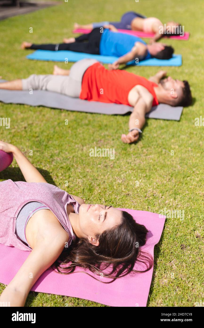 Giovane donna sdraiata sul materassino per esercizi con gli occhi chiusi mentre pratica lo yoga con le persone nel parco Foto Stock